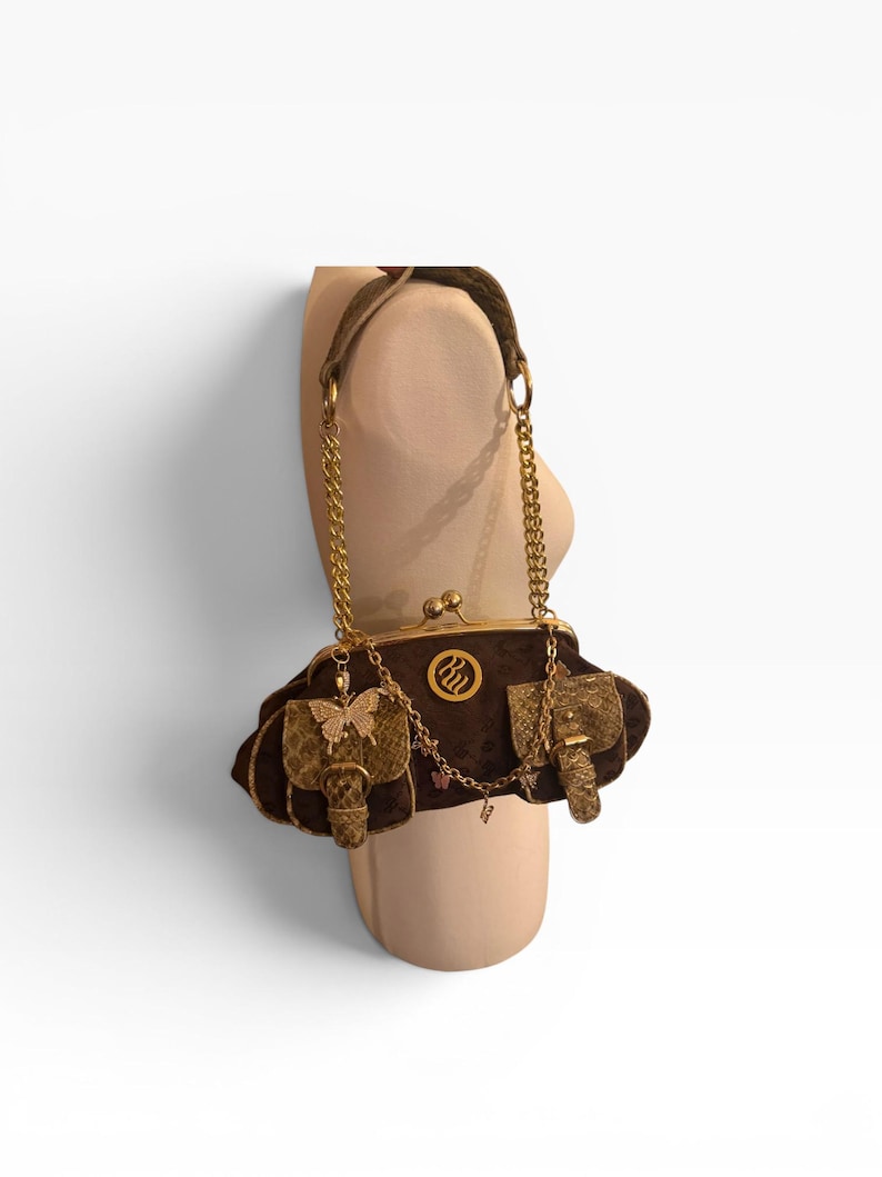 May include: A brown velvet handbag with gold chain straps and accents. The bag features gold butterfly and elephant embellishments, a gold clasp, and a decorative logo. The bag is displayed on a mannequin.