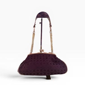 May include: A vintage dark purple handbag with a gold-coloured clasp and chain straps. The bag has a patterned design and is displayed on a stand. The bag's frame and chain straps are gold-coloured.