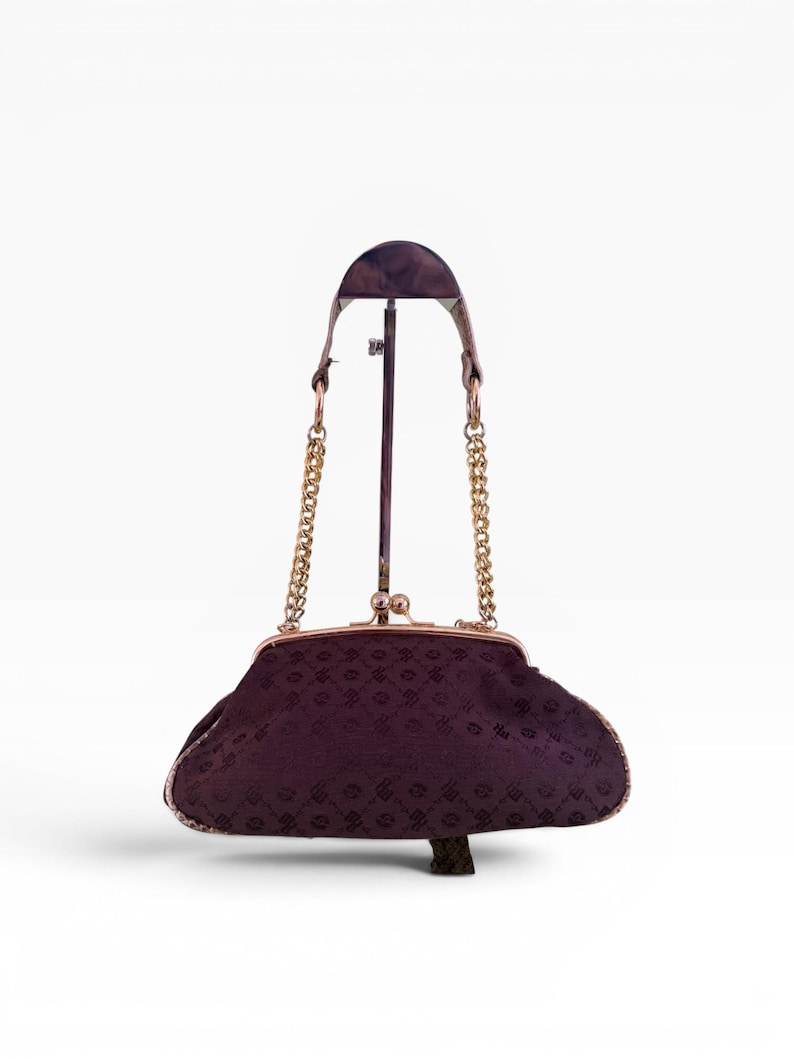 May include: A vintage dark purple handbag with a gold-coloured clasp and chain straps. The bag has a patterned design and is displayed on a stand. The bag's frame and chain straps are gold-coloured.