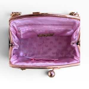 May include: An open black handbag with a gold-coloured chain strap and clasp. The interior is lined with pink satin fabric, featuring a small zipped pocket and a decorative label. The bag has a kiss-lock closure.