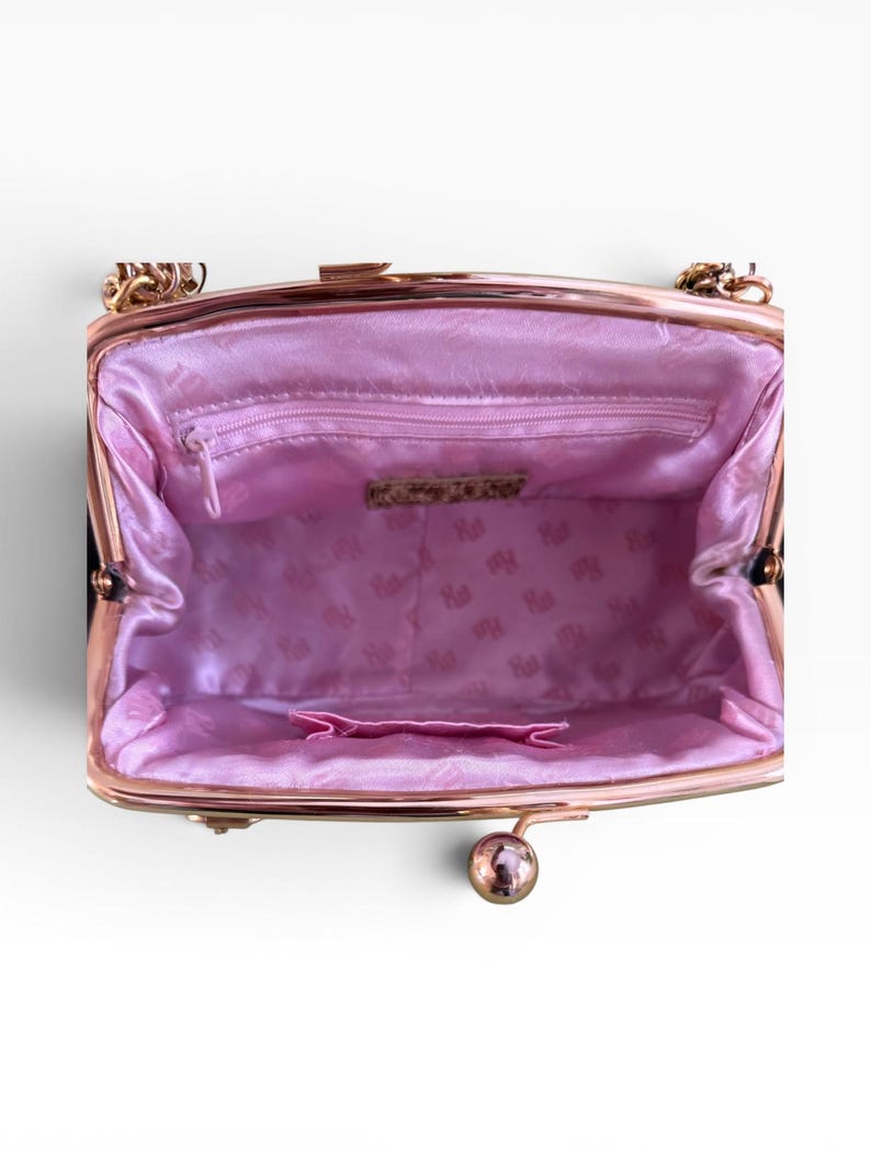 May include: An open black handbag with a gold-coloured chain strap and clasp. The interior is lined with pink satin fabric, featuring a small zipped pocket and a decorative label. The bag has a kiss-lock closure.