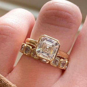 May include: A gold ring with a large, square-cut diamond is the focal point, complemented by a smaller band with multiple smaller stones. The rings are set against a neutral background, highlighting the jewelry's sparkle and design.