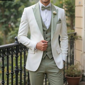 Puede incluir: Un traje formal en color marfil con detalles en verde salvia. El conjunto incluye una chaqueta con solapa de satén, un chaleco y un pantalón. Una pajarita y un pañuelo de bolsillo a juego completan el conjunto. Una flor blanca está prendida en la solapa.