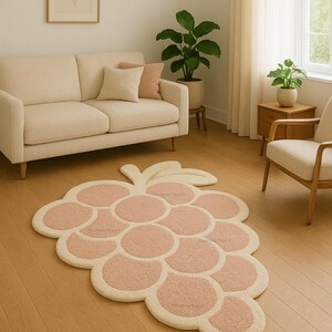 May include: A novelty rug in the shape of a bunch of grapes. The rug has a light pink design for the grape clusters, with a cream-coloured border and stem. It is placed on a wooden floor in a living room.