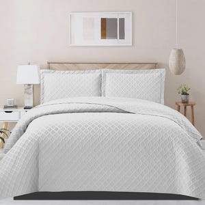 May include: White bedspread and two matching pillowcases with a textured, geometric pattern. The bedding is on a wooden bed frame, with a lamp and artwork visible in the background. The overall theme is bedroom decor.
