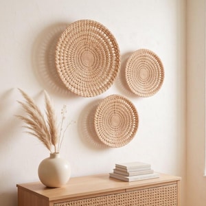 May include: Three circular woven wall baskets made of light tan natural fibres, displayed in a cluster on a wall. The baskets feature a concentric coil pattern with an open weave design, varying in size from one large to two smaller pieces.