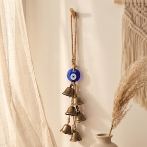 Può includere: Una decorazione da parete composta da una perla in vetro blu a forma di occhio protettivo, sospesa a una spessa corda di iuta. Sotto la perla, sei piccoli campanelli in metallo color ottone anticato sono fissati alla corda in una disposizione a cascata.