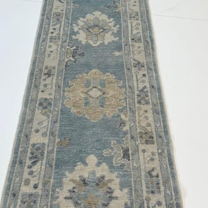 Könnte beinhalten: Ein langer, rechteckiger Teppich mit einem zentralen türkisfarbenen Feld und komplizierten Blumenmustern in Beige und Grau. Der Teppich hat einen Rand mit sich wiederholenden geometrischen Designs. Der Teppich ist für einen Flur oder Eingangsbereich konzipiert.
