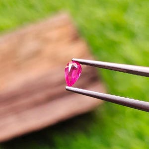 May include: A pear-shaped, faceted gemstone with a vibrant, saturated pinkish-red color. The gemstone is held securely by a pair of silver-toned metal tweezers, showcasing its clear, reflective surface and precise, symmetrical cut.
