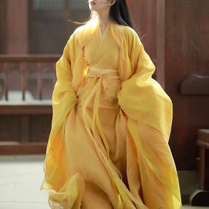May include: A flowing, yellow traditional dress with long sleeves and a matching sash. The dress appears to be made of a lightweight, sheer fabric. The person is standing in a wooden structure with natural light.