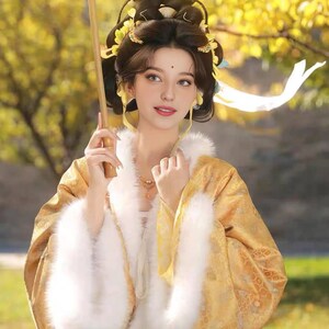 May include: A woman in traditional Asian dress, holding a yellow parasol. The outfit is gold with white fur trim. Her hair is styled with ornate decorations. The background features yellow foliage, suggesting an outdoor setting.