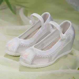 May include: A pair of white satin wedge shoes with delicate floral embroidery on the toe and a scalloped lace trim. The shoes have a strap across the top and a woven insole, perfect for special occasions.