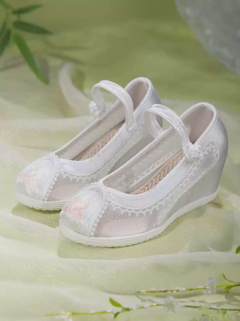 May include: A pair of white satin wedge shoes with delicate floral embroidery on the toe and a scalloped lace trim. The shoes have a strap across the top and a woven insole, perfect for special occasions.