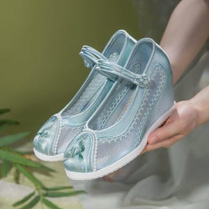 May include: A pair of light blue wedge shoes with a floral design on the toe. The shoes have a strap across the top and a white sole. The shoes are made of a sheer, lacy material.