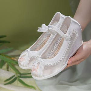 May include: A pair of white wedge-heeled shoes with delicate floral embroidery on the toe and sides. The shoes feature a strap across the top and are made with a combination of satin and lace, creating an elegant and feminine look.
