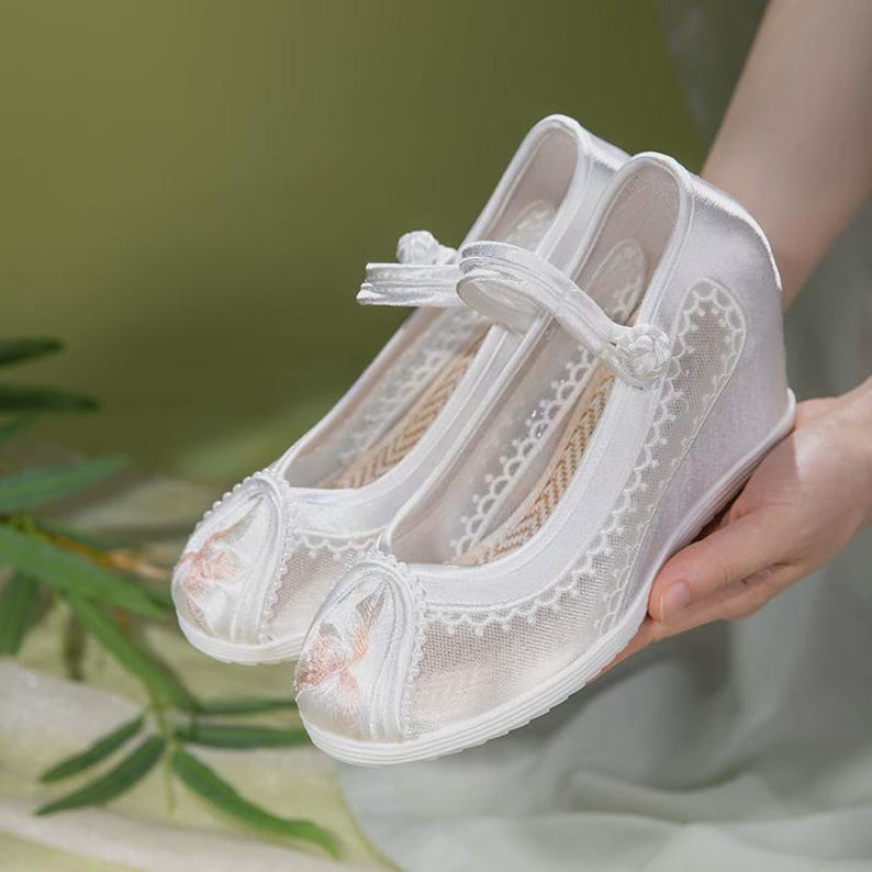 May include: A pair of white wedge-heeled shoes with delicate floral embroidery on the toe and sides. The shoes feature a strap across the top and are made with a combination of satin and lace, creating an elegant and feminine look.