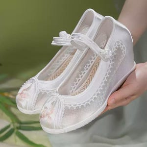 May include: A pair of white satin shoes with a wedge heel. The shoes feature a delicate floral embroidery on the toe and a lace overlay. A strap with a small bow secures the shoe. The shoes are held by a person.