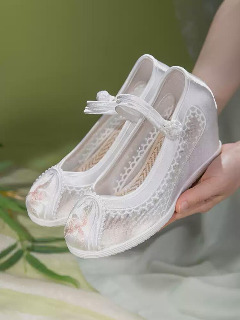 May include: A pair of white satin shoes with a wedge heel. The shoes feature a delicate floral embroidery on the toe and a lace overlay. A strap with a small bow secures the shoe. The shoes are held by a person.