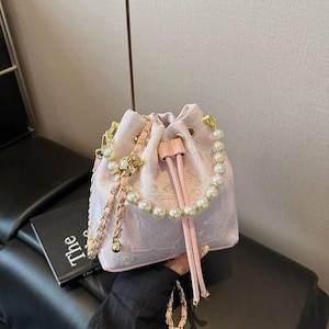 Puede incluir: Un bolso tipo cubo rosa claro con cordón y estampado floral. El bolso presenta una correa de cadena dorada y perlas, un ribete con cuentas de perlas y cordones rosas. Fabricado en tejido suave y texturizado.