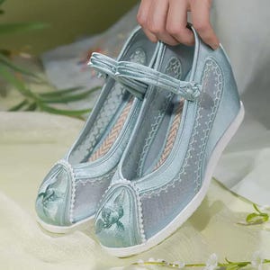 May include: A pair of light blue wedge shoes with a floral design on the toe. The shoes have a mesh overlay and a strap across the top. The soles are white, and the shoes are held up by a hand.