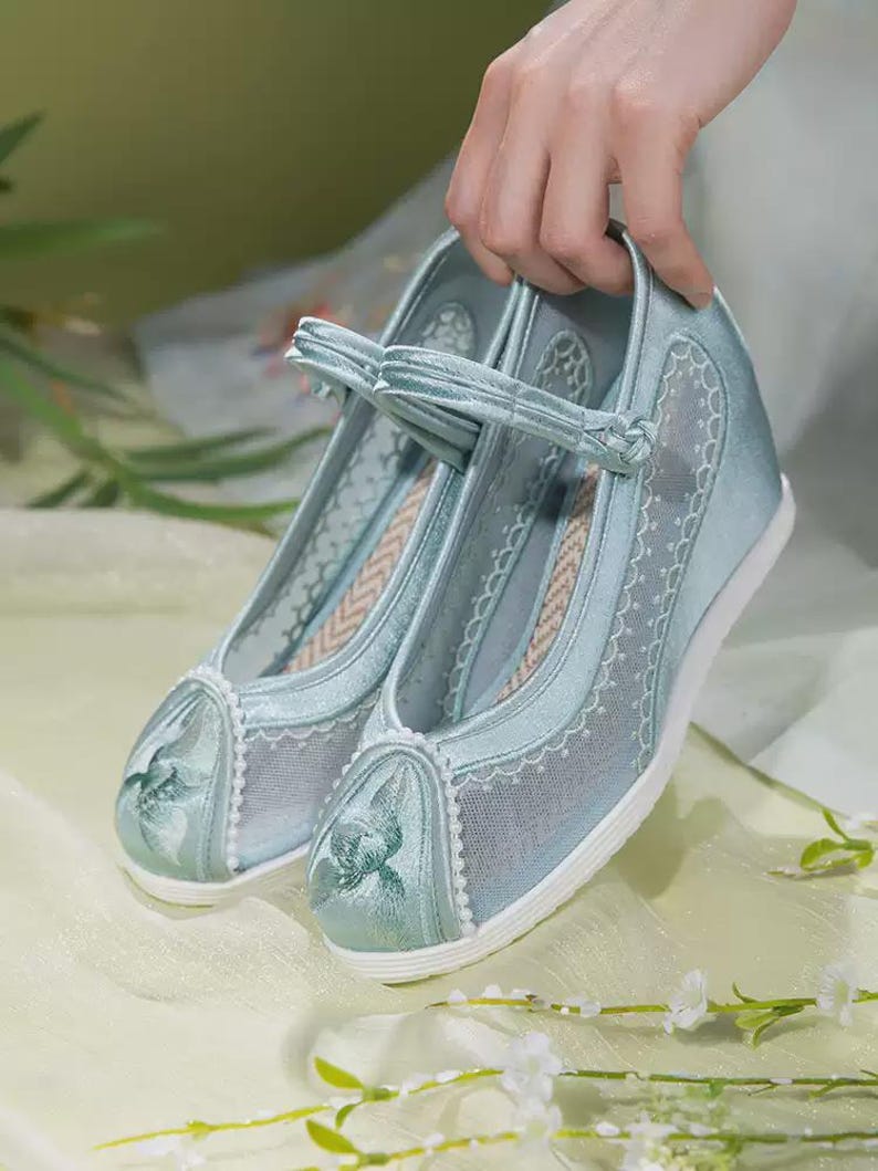 May include: A pair of light blue wedge shoes with a floral design on the toe. The shoes have a mesh overlay and a strap across the top. The soles are white, and the shoes are held up by a hand.