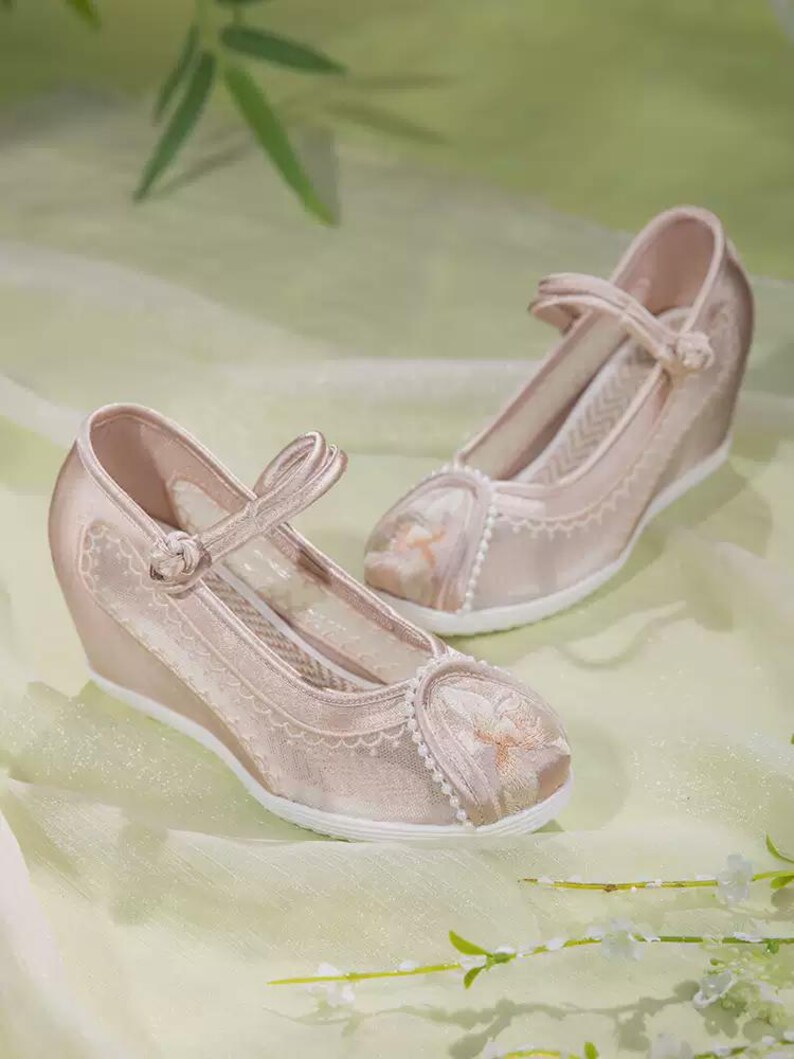 May include: A pair of blush-colored wedge heels with intricate embroidery and delicate straps. The shoes feature a closed-toe design with floral embroidery and a white sole. The heels are approximately 3 inches high.