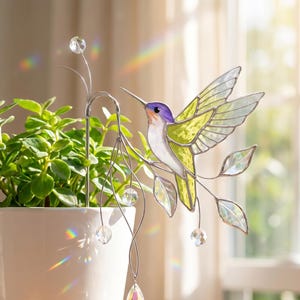 May include: A stained glass hummingbird with green and yellow wings, a purple head, and a long beak perches on a metal branch with crystal accents. The decorative piece is next to a potted plant in a white pot.