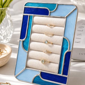May include: A rectangular ring display stand featuring a stained glass frame with shades of light blue, teal, and dark blue glass pieces joined by silver metal solder. The center contains five horizontal rows of cream-colored velvet ring slots.