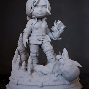 Peut inclure: Figurine grise imprimée en 3D d'une guerrière debout sur une base rocheuse, tenant un livre en flammes. La figurine porte une armure et des bottes. La base représente un dragon vaincu. Le texte "EDYTHE CROSS 3D" est dans le coin supérieur gauche.