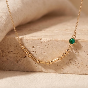 May include: A gold chain necklace featuring a series of small gold beads and cylindrical spacers arranged in a sequence, accented by a single round, bezel-set green gemstone positioned asymmetrically along the delicate metal link chain.