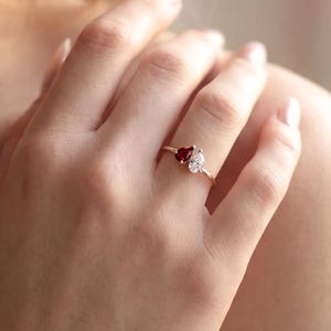 May include: A gold ring featuring a heart-shaped red gemstone and a pear-shaped white gemstone. The ring is worn on a person's finger. The ring's band is thin and delicate.