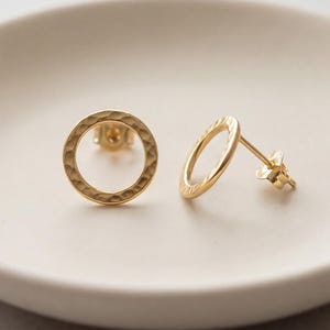 May include: A pair of gold-colored stud earrings, each featuring an open circle design with a textured surface. The earrings are displayed on a white, slightly curved surface, showcasing their simple yet elegant style. The earrings are approximately 0.75 inches in diameter.