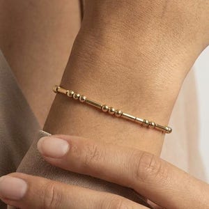 May include: A delicate gold bracelet featuring a series of small, evenly spaced beads and cylindrical bars. The bracelet is worn on a wrist, with the gold color contrasting against the skin tone. The jewelry piece is simple and elegant.