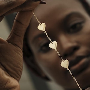 May include: A gold-colored necklace featuring three heart-shaped pendants. Each heart is engraved with a name: Elizabeth, Sophia, and Mekenzie. The necklace is held up against a blurred background.