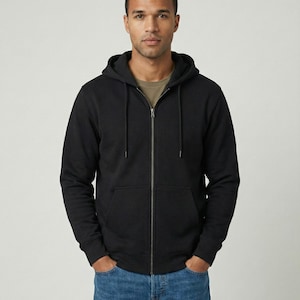 May include: A black zip-up hoodie featuring a drawstring hood, long sleeves, and a front kangaroo pocket. The garment is worn over a plain olive green t-shirt and paired with blue denim jeans, showcasing a standard fit and a full-length metal zip fastener.
