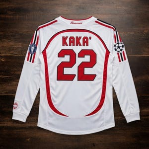 Puede incluir: Camiseta de fútbol blanca de manga larga con detalles rojos y negros. La camiseta muestra el nombre "KAKA'" y el número "22" en rojo en la espalda. Incluye un gráfico de balón de fútbol y la palabra "Rossoneri" en el cuello.