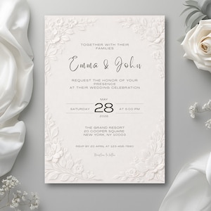 Può includere: Un invito a nozze color crema con dettagli floreali in rilievo e una scrittura elegante. L'invito recita "Emma & John" e include la data, l'ora e il luogo del matrimonio. Una rosa bianca e tessuto drappeggiato sono sullo sfondo.