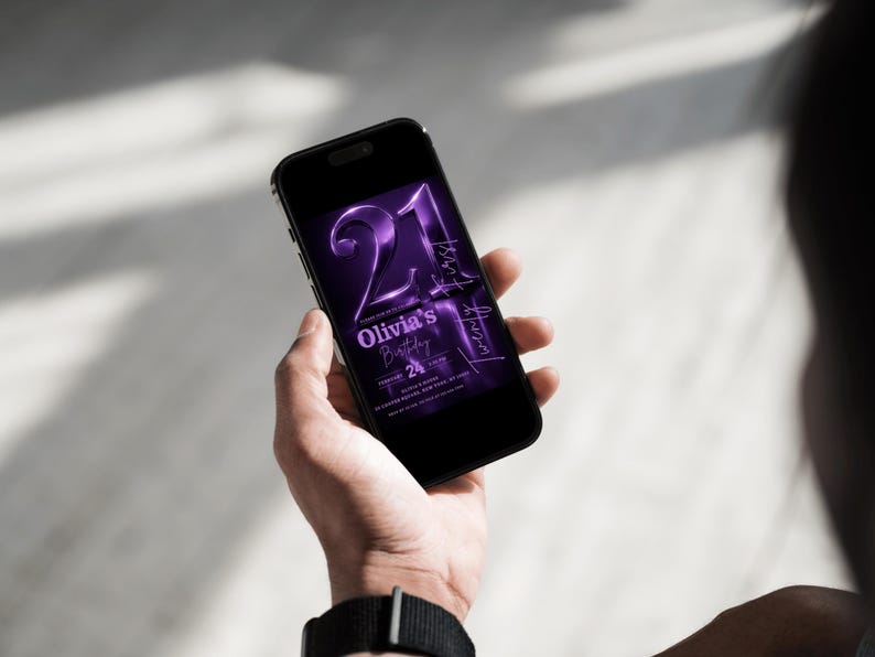 May include: A black smartphone displays a purple digital invitation for "Olivia's Birthday." The invitation features the number "21" in a bold, metallic font, with additional text indicating the date and time of the event. The phone is held in a person's hand.