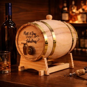 Puede incluir: Un barril de madera con bandas de latón y un grifo, apoyado sobre un soporte de madera. El barril está grabado con la frase "Not a Day Over Fabulous". Una botella y un vaso de líquido están en el fondo.