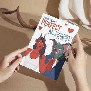 May include: A greeting card with the text "I THINK WE HAVE PERFECT SYNERGY" and illustrations of a red-skinned demon and a green bat-like creature. The card has red heart accents and is held by a person.