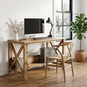 Pode incluir: Uma secretária de madeira com design lateral em X e prateleira inferior, acompanhada por uma cadeira de madeira clara com assento entrançado. Na secretária encontram-se um monitor de computador, um candeeiro preto ajustável e um vaso branco.
