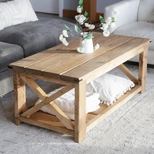May include: A rectangular wooden coffee table with an X-shaped design on the sides. The table has a lower shelf and is made of light-colored wood. Two white pillows are on the shelf. A vase of white flowers sits on top.