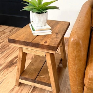 May include: A rustic, wooden side table with a lower shelf. The table is made of light brown wood and has a natural finish. A small, white pot with a green plant and a stack of books sits on top. The table is approximately 24 inches tall.