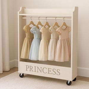Puede incluir: Un perchero crema para ropa infantil con cinco vestidos en tonos amarillo, azul y rosa. El perchero tiene un estante y la palabra "PRINCESS" en letras grises. El perchero está sobre ruedas.