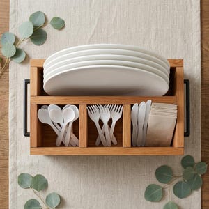 Può includere: Un contenitore rettangolare in legno con manici in metallo nero contenente una pila di piatti bianchi nella sezione superiore e tre scomparti inferiori con cucchiai, forchette e coltelli in plastica bianca e un pacchetto di tovaglioli beige.
