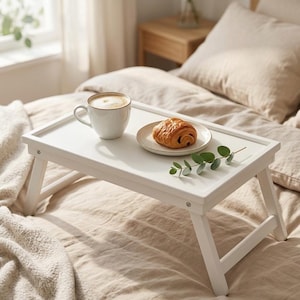 May include: A white wooden breakfast tray with folding legs, holding a cup of coffee and a croissant on a small plate. A sprig of greenery adds a touch of freshness. The tray is set on a bed with beige bedding.