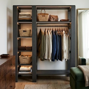 Op de afbeelding: Een open kastsysteem van donker hout met geïntegreerde verlichting, voorzien van verticale planken met gevlochten manden en opgevouwen kleding, naast een kledingroede met diverse blazers, overhemden en broeken op houten hangers.