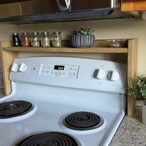 Puede incluir: Un especiero de madera se encuentra sobre una estufa eléctrica blanca. El estante contiene pequeños frascos de vidrio con especias, una suculenta en maceta y un cuenco pequeño. El panel de control de la estufa muestra la hora.