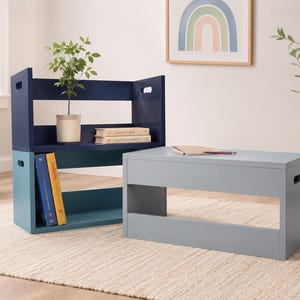 Può includere: Un set di tre cassette rettangolari in legno con maniglie intagliate. Una cassetta è blu scuro, una verde acqua e una grigio chiaro. Le cassette sono impilate e disposte per fungere da scaffale modulare e da panca bassa.