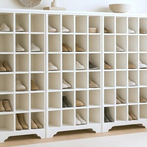 Op de afbeelding: Een witte schoenenkast met meerdere vakken, elk met een paar schoenen. De schoenen zijn in verschillende neutrale kleuren. Links staat een decoratieve plant in een witte pot. Er ligt een neutraal tapijt voor de kast.